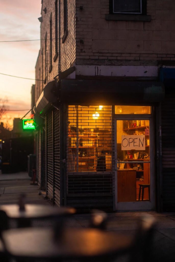 A slightly gritty but hopeful photo of a small coffee shop in Queens at sunrise. Metal gate halfway up. Warm light spilling out.
